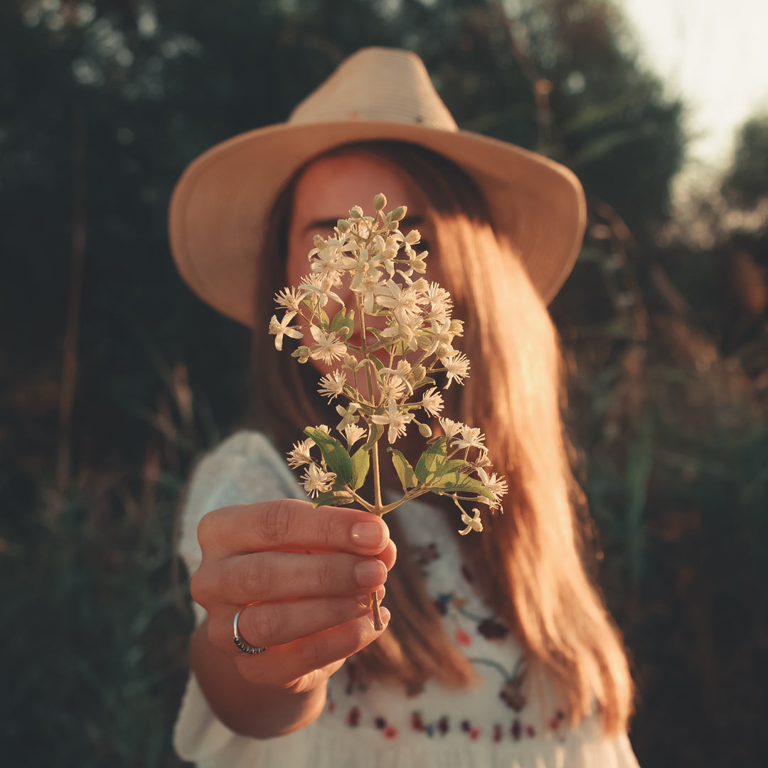 A woman wearing a straw hat and a white embroidered dress holds a delicate branch of small white flowers towards the camera.