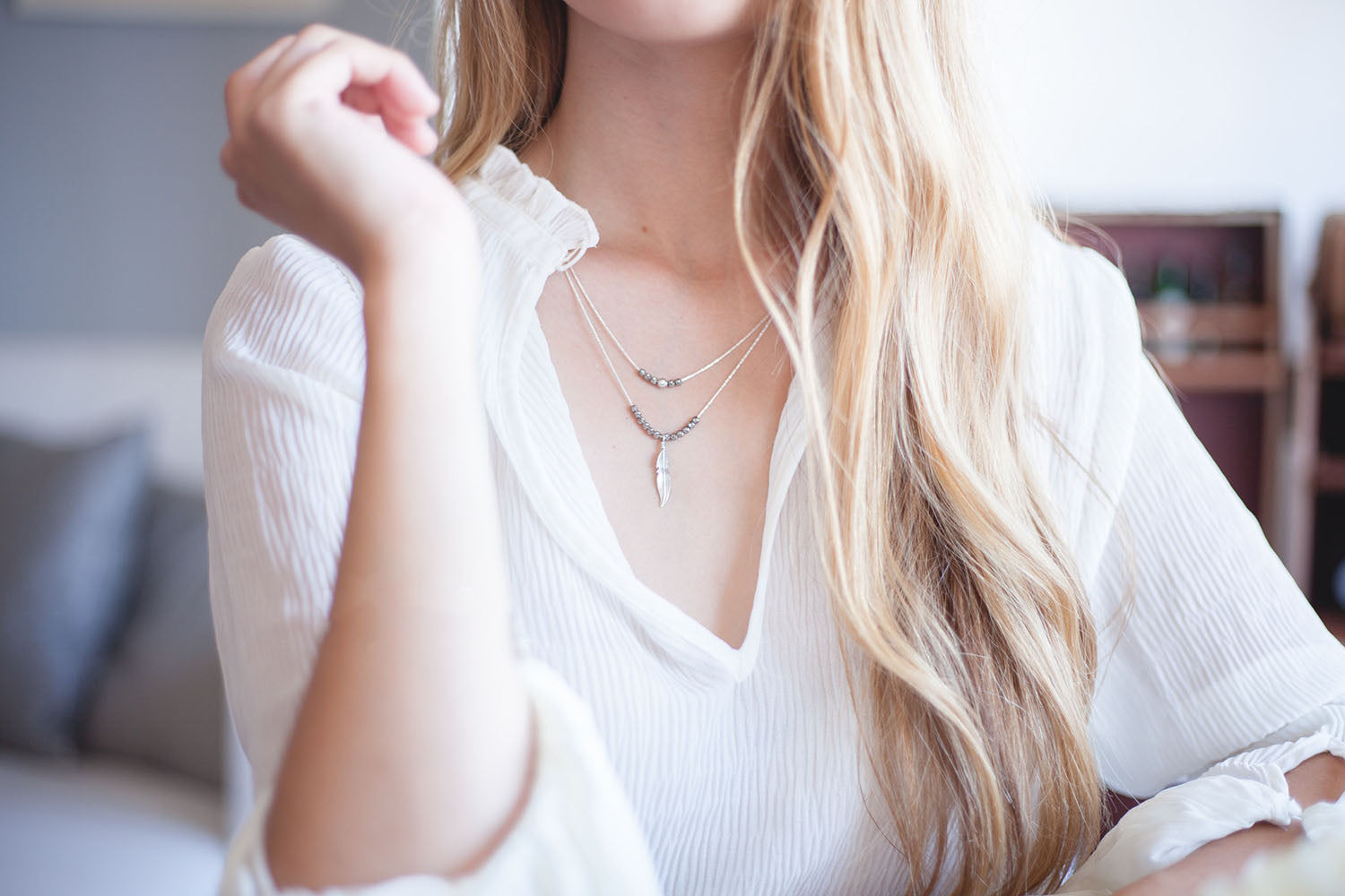 A woman with long blonde hair wearing a delicate layered silver necklace with a feather pendant, dressed in a white blouse, sitting in a softly lit indoor setting.