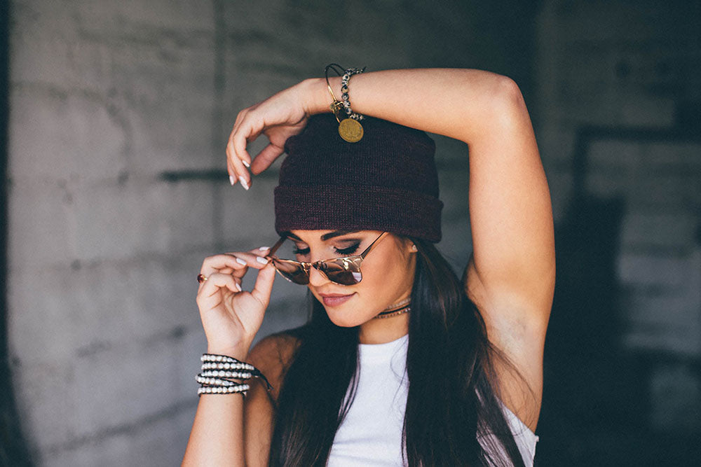 A stylish woman wearing a dark beanie, sunglasses, and layered jewelry, adjusting her shades with a confident expression. She sports a white tank top, accessorized with bracelets and a coin charm bracelet, against an urban, moody backdrop.