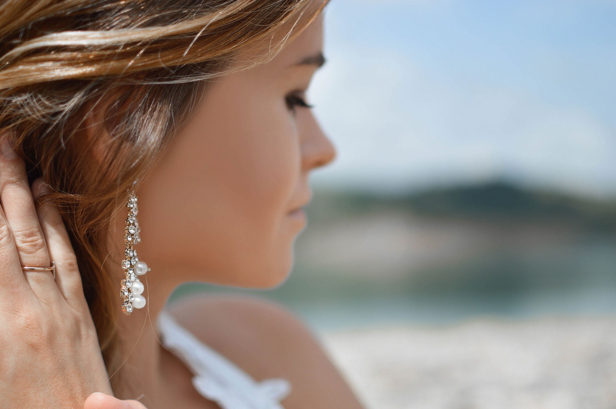 A woman with sun-kissed skin and light brown hair wearing an elegant silver earring adorned with pearls and crystals, gazing into the distance with a blurred ocean and sky in the background.
