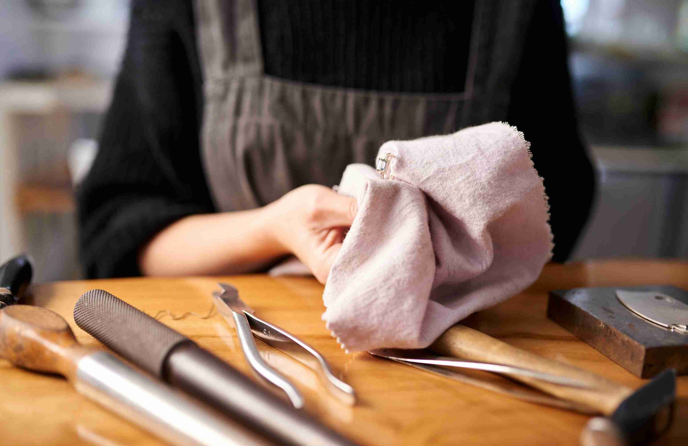 A jeweler carefully polishing a silver ring with a soft cloth, surrounded by professional jewelry-making tools on a wooden workbench. The artisan is wearing a dark sweater and an apron, focusing on craftsmanship and detail.