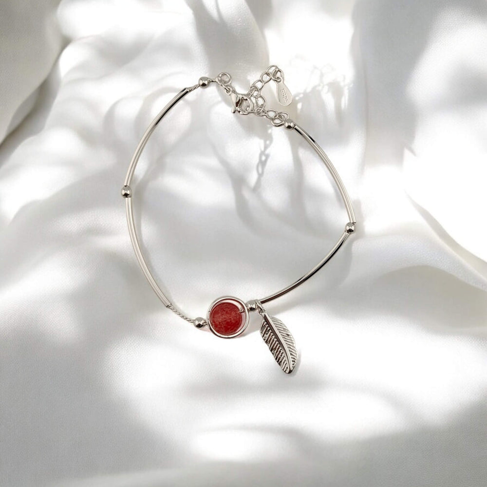 Silver bracelet with red charm and feather pendant laid on white fabric