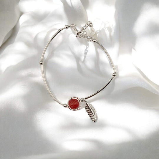 Silver bracelet with red charm and feather pendant laid on white fabric